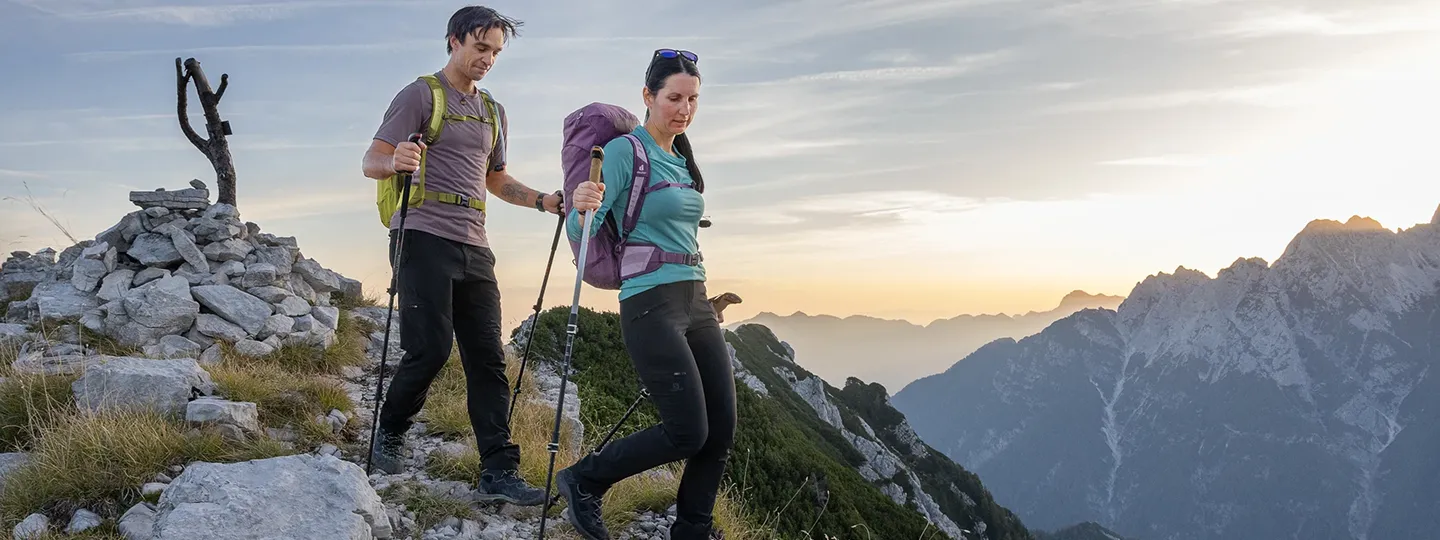 Zwei Wanderer mit Rucksäcken und Stöcken auf Bergpfad im Sonnenuntergang, im Hintergrund ein Panorama mit Alpen-Gipfeln.
